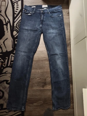 Blå raka jeans från Calvin Klein - Säljer ett par klassiska blå jeans från Calvin Klein med rak passform och fem fickor. Jeansen har subtila slitningar och tydliga sömmar, samt CK-logga på bakfickan. Perfekta för en avslappnad och stilren look.