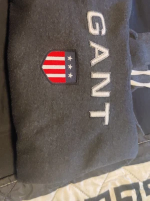 Mörkgrå hoodie från GANT - Snygg mörkgrå hoodie från GANT med broderad logga och amerikanskinspirerat sköldmärke på bröstet. Klassisk känguruficka framtill och vita dragsnören i huvan. Perfekt för en avslappnad och sportig stil.