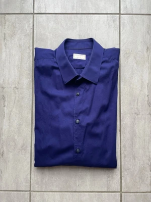 Blå/lila skjorta, tiger of sweden  - Stilren blå/lila skjorta från Tiger of sweden i klassisk modell. Skjortan har knappar framtill, lång ärm med manschett och en snygg krage. Perfekt för dig som gillar minimalistisk och tidlös stil. Tillverkad i mjuk bomull för skön känsla. Vid frågor och funderingar skriv gärna! 