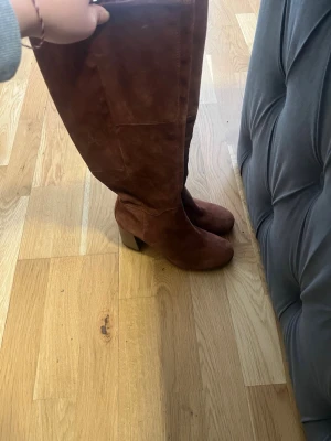 Bruna höga mockastövlar med klack - Säljer ett par snygga bruna stövlar i mocka med hög skaft och bred klack. Stövlarna har en stilren design och passar perfekt till höst- och vinteroutfits. De är utan synligt märke och har en mjuk, matt finish.