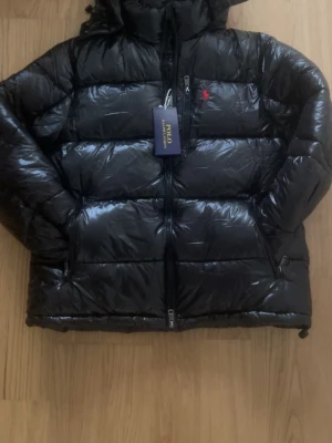 Svart dunjacka från Polo Ralph Lauren - Snygg svart dunjacka från Polo Ralph Lauren med glansig finish och röd logga på bröstet. Jackan har hög krage, dragkedja framtill och två sidofickor med dragkedja. Perfekt för kalla vinterdagar och riktigt stilren look.