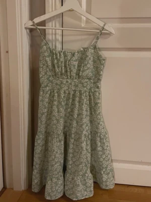 Grön blommig sommarklänning - Söt ljusgrön klänning med vita blommor och smala axelband. Klänningen har smock i midjan och volangig kjol, perfekt för varma dagar. Lätt och luftig modell som ger en avslappnad känsla.