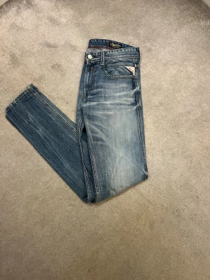 Replay Anbass - Snygga blå slim fit jeans från Diesel med klassisk femficksdesign och diskreta slitningar. Jeansen har en ljus tvätt och detaljerade sömmar, samt en liten logotyp på myntfickan. Perfekta för en avslappnad och trendig look.