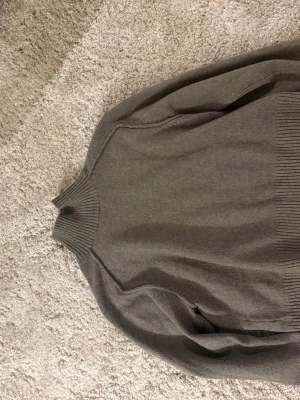 Grå stickad tröja med hög krage - En stilren grå stickad tröja med hög ribbad krage och ribbade muddar. Tröjan har en enkel design och är perfekt för lager-på-lager. Materialet känns mjukt och bekvämt, och modellen är rak med långa ärmar.