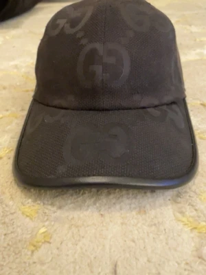 Svart keps från Gucci med GG-mönster - Snygg svart keps från Gucci med det klassiska GG-monogrammet ton-i-ton över hela kepsen. Kepsen har böjd skärm och är tillverkad i ett textilmaterial med läderdetalj längs skärmens kant. Perfekt för en trendig och exklusiv look.