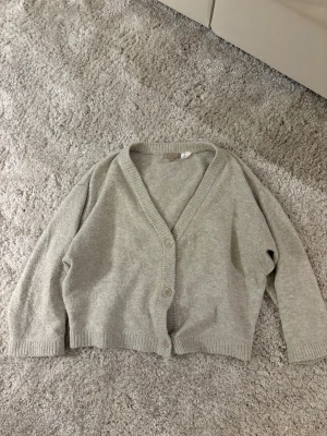 Beige kofta från H&M - Mysig ljusgrå kofta från H&M med v-ringning och två knappar framtill. Koftan har ribbade muddar vid ärmslut och nederkant, samt en avslappnad passform. Perfekt att slänga över en t-shirt för en chill look. Lite nopprig.