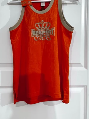 Orange linne från Adidas Respect ME - Snyggt orange linne från Adidas Respect ME-kollektionen (Missy Elliot) med guldfärgade detaljer och tryck. Linnet har breda beige muddar och ett mönster med kronor i tyget. Materialet är glansigt och lätt, perfekt för en sportig och trendig look.