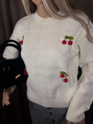 Vit stickad tröja med körsbärsdetaljer - Söt vit stickad tröja med broderade körsbär i rött och grönt på bröstet och sidan. Tröjan har rund halsringning, långa ärmar och ribbade muddar. Perfekt för dig som gillar lekfulla detaljer och vill sticka ut lite extra.