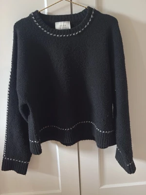 Svart stickad tröja med vita detaljer - Mysig svart stickad tröja med vita sömmar längs halsringning, ärmslut och nederkant. Tröjan har en avslappnad passform, rund hals och långa ärmar. Perfekt för kyliga dagar och enkel att matcha till olika outfits.
