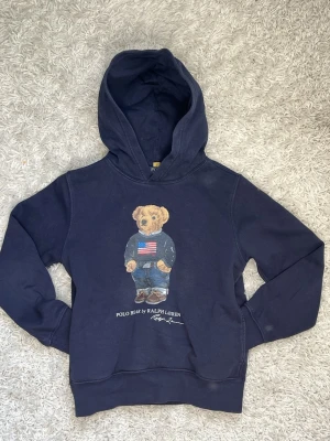 Mörkblå hoodie Polo Bear Ralph Lauren - Mörkblå hoodie från Ralph Lauren med ikoniskt Polo Bear-tryck på bröstet. Björnen har på sig jeans, boots och en tröja med amerikanska flaggan. Hoodien har huva och magficka. Perfekt för dig som gillar klassisk streetstyle och snygga detaljer.
