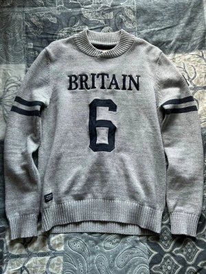 Grå stickad morris tröja - Grå stickad tröja från morris med svart text 'BRITAIN' och siffran 6 framtill. Två svarta ränder på ärmarna och ribbstickade muddar vid hals, ärmslut och nederkant. Perfekt för en sportig och avslappnad stil.