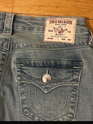 True Religion ljusblå bootcut jeans - Säljer ett par ljusblå jeans från True Religion med bootcut passform och ikoniska ficklock bak med kontrastsömmar och silverfärgade knappar. Klassisk denimlook i bomull, perfekt för dig som gillar streetwear och retrostil.