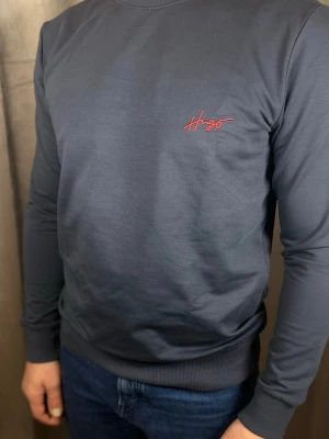 Mörkblå stickad tröja från Hugo - Mörkblå stickad tröja från Hugo med röd broderad logga på bröstet. Tröjan har rund halsringning, långa ärmar och ribbade muddar vid ärmslut och nederkant. Perfekt för en stilren och enkel look.