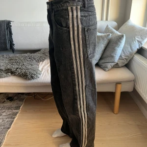 Svarta Adidas jeans med ränder - Säljer ett par svarta raka jeans från Adidas med de klassiska vita ränderna längs sidorna. Jeansen har hög midja, fem fickor och är tillverkade i denim. Perfekta för en sportig och avslappnad stil.