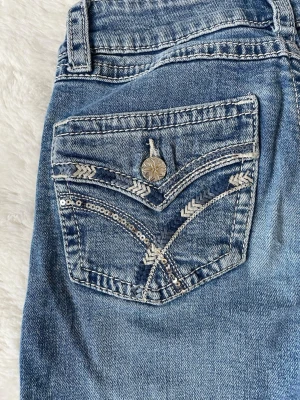 Blå bootcut jeans med paljettdetaljer - Säljer ett par blå bootcut jeans med snygga paljettdetaljer och mönster på bakfickan från Gina Young