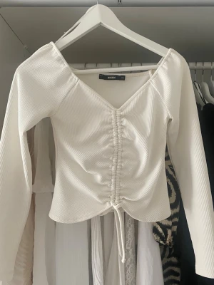Vit ribbad topp med snörning Bik Bok - Trendig vit ribbad topp från Bik Bok med lång ärm och v-ringning. Toppen har snygg snörning framtill som ger en rynkad effekt och en feminin siluett. Perfekt att styla med jeans eller kjol för en cool look.