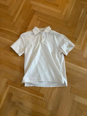 Vit pikétröja med krage - Klassisk vit pikétröja med kort ärm och krage. Tröjan har knäppning med tre knappar framtill och är tillverkad i ett mjukt, strukturerat material. Perfekt för en clean och enkel stil.