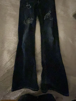 Mörkblå bootcut jeans med slitningar - Säljer ett par mörkblå bootcut jeans med slitna detaljer på låren och knäna. Jeansen har utsvängda ben och klassisk femficksdesign. Perfekta för dig som gillar en avslappnad och trendig look.