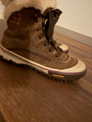 MERRELL Bruna kängor med foder och snörning - Bruna kängor med mjukt vitt foder och grov snörning. Ovandel i mockaliknande material och detaljer i rutigt tyg vid skaftet. Tjock sula med vit och lila kant. Perfekta för kalla dagar och riktigt sköna att ha på sig.