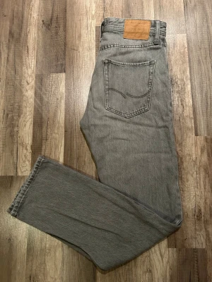 Grå raka jeans från Jack & Jones  - Snygga grå jeans från Jack & Jones med rak passform och klassisk femficksdesign. Jeansen har en diskret logga på bakfickan. Tillverkade i slitstark bomull, perfekta för en clean och avslappnad stil. Storlek W28 L32