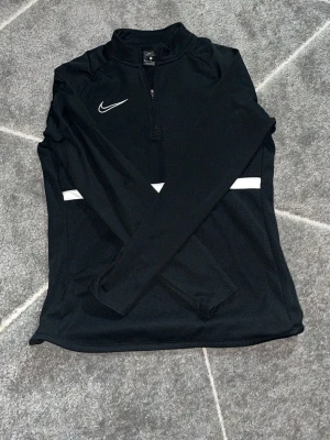 Svart långärmad träningströja Nike - Svart långärmad träningströja från Nike med halv dragkedja framtill och vit logga på bröstet. Tröjan har en vit rand över bröstet och är tillverkad i ett stretchigt funktionsmaterial som passar perfekt för träning.