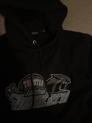 Svart Trapstar Shooters hoodie - Säljer en svart hoodie från Trapstar med stor grå och vit Shooters-logga och pantermotiv på bröstet. Klassisk luva med mjukt material, perfekt för en avslappnad streetwear-look. Hoodien har långa ärmar och ribbade muddar.
