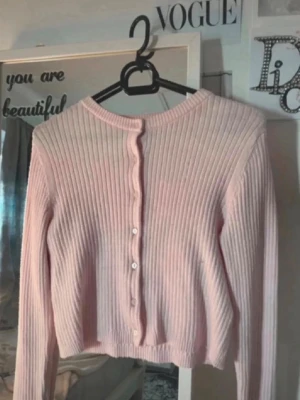 Ljusrosa ribbad kofta med knappar - Supersöt ljusrosa kofta med ribbad struktur och rund hals. Koftan har långa ärmar och knäpps framtill med små vita knappar. Perfekt att slänga över en topp för en chill och trendig look.