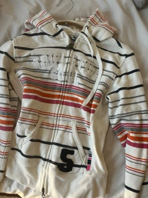 Redan sold - Vit hoodie med dragkedja från Triple Five Soul, dekorerad med broderad text på bröstet och en stor svart siffra 5 på fickan. Hoodien har färgglada ränder i svart, rött, orange och rosa samt en stor huva och fickor framtill. Perfekt för en avslappnad streetwear-look.