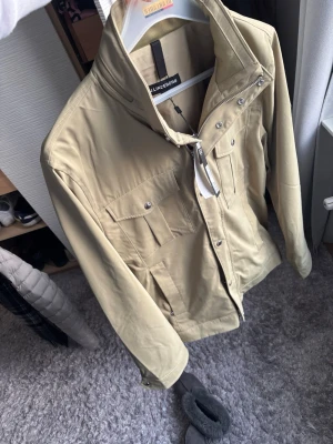 Vår jacka från J.Lindeberg - Stilren olivgrön jacka från J.Lindeberg med hög krage, dragkedja och tryckknappar. Jackan har flera praktiska fickor framtill och en modern, rak passform. Perfekt för dig som gillar minimalistisk och snygg design.