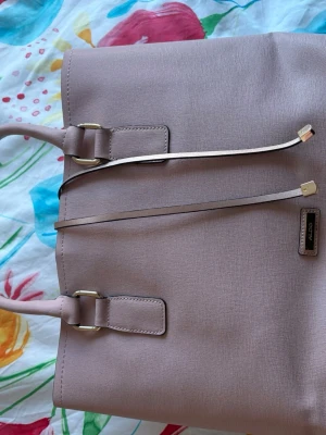 Ljusrosa handväska från ALDO - Stilren ljusrosa handväska från ALDO med två handtag och dekorativa guldfärgade detaljer. Väskan har en fyrkantig form och är tillverkad i ett strukturerat konstläder. Perfekt för dig som gillar klassisk och enkel design.