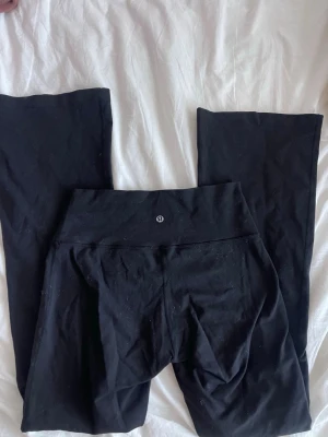 Svarta bootcut leggings från Lululemon - Säljer ett par svarta leggings från Lululemon med bootcut-ben och hög midja. De är tillverkade i ett stretchigt och mjukt material som sitter skönt på kroppen. Perfekta för dig som gillar en enkel men snygg look.