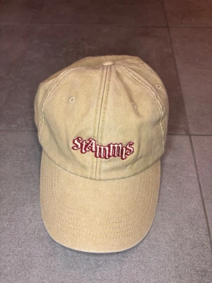 Beige keps från Spotify x Stamms - Säljer en beige keps i bomull med broderad text 'Stamms' i rött framtill och Spotify-logga på sidan. Klassisk dad cap-modell med justerbart spänne bak. Perfekt accessoar för en avslappnad streetwear-look.