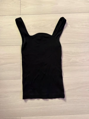 Svart ribbad linnetopp - En enkel och stilren svart ribbad linnetopp med breda axelband. Toppen är ärmlös och har en tight passform som framhäver figuren. Perfekt att matcha med jeans eller kjol för en clean look.