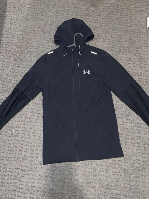 Svart vindjacka från Under Armour - Svart vindjacka med huva från Under Armour. Jackan har hel dragkedja, reflekterande detaljer på axlarna och logga på bröstet. Lätt material och sportig passform, perfekt för blåsiga dagar och träning utomhus.
