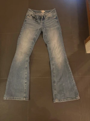 Gina Young 14+ jeans - Säljer ett par ljusblå bootcut jeans med snygg slitning och broderade bakfickor. Jeansen har klassisk femficksmodell och vida ben. Perfekta för dig som gillar retrostil och vill ha något som sticker ut lite extra.