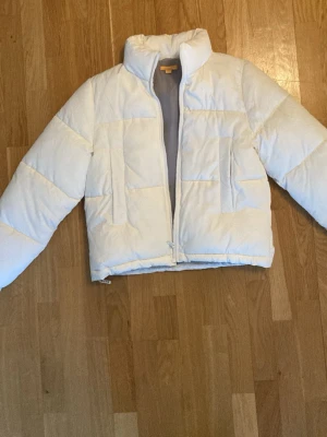 Vit pufferjacka från Gina Tricot - Snygg vit pufferjacka från Gina Tricot med hög krage och dragkedja framtill. Jackan har en boxig passform och quiltade paneler som ger en trendig look. Perfekt för kalla dagar och enkel att matcha med olika outfits.