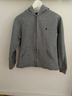 Äkta Grå hoodie med dragkedja Polo Ralph Lauren  - Äkta Grå hoodie från Polo Ralph Lauren med dragkedja, huva och två stora fickor framtill. Klassisk design med liten broderad logga på bröstet. Tillverkad i mjukt sweatshirtmaterial som känns skönt mot huden.