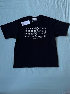 Svart Maison Margiela t-shirt - Svart t-shirt från Maison Margiela med ikoniskt tryck framtill i vitt med siffror och logga. Klassisk passform och rund hals. Perfekt för dig som gillar stilrena och trendiga plagg med designer-vibe.