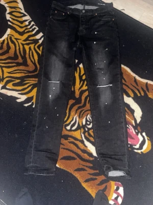 Svarta jeans från Purple Brand - Svarta jeans från Purple Brand med slitningar på knäna och vita färgstänk. Klassisk femficksmodell med raka ben och knappgylf. Jeansen har en cool, streetinspirerad look och är tillverkade i denim.