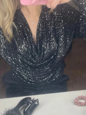 Glittrig svart topp med drapering - Säljer en svart långärmad topp med silvriga glitterdetaljer och snygg drapering framtill. Toppen har djup v-ringning och dekorativa knappar i guld på ärmsluten. Perfekt för dig som vill sticka ut med en festlig look.