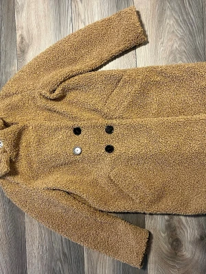 Brun teddyjacka från Zara  - Mysig brun teddyjacka från Zara med dubbelknäppning och svarta knappar. Jackan har en längre modell, två sidfickor och är fodrad med svart tyg på insidan. Perfekt för kalla dagar när du vill vara både varm och trendig.