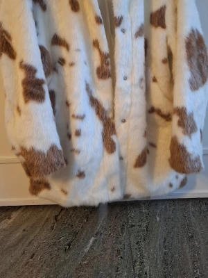 Vit och brun fuskpälsjacka - Säljer en fluffig fuskpälsjacka i vitt med bruna fläckar. Jackan har knappar framtill och en oversized passform som ger en cool och trendig look. Perfekt för dig som vill sticka ut i vinter och hålla dig varm med stil.