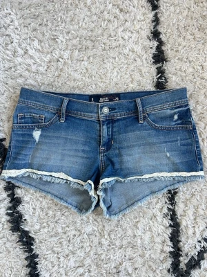 Hollister Shorts 💙 - Snygga blå jeansshorts från Hollister i storlek w29, short short med låg midja, slitna detaljer och råa kanter. Shortsen har vit spets längs bensluten och klassiska fickor både fram och bak. Midjemått 43cm mätt rakt över 💙