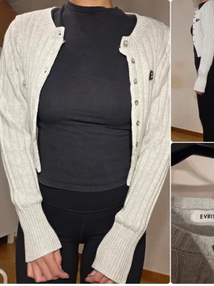 Ljusgrå ribbad kofta från EVRIS - Superfin ljusgrå kofta från EVRIS med ribbad struktur och knappar framtill. Croppad modell med långa ärmar och liten broderad detalj på bröstet. Perfekt att slänga över en topp för en chill look.