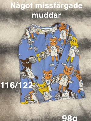 Blå långärmad  t-shirt med kattmönster Mini Rodini - Blå långärmad t-shirt från Mini Rodini med färgglatt kattmönster där katterna är utklädda till cheerleaders. T-shirten har långa  ärmar och knappar vid halsen. Perfekt för dig som gillar lekfulla prints och vill sticka ut.🌸 Något missfärgade muddar. Storlek 116/122