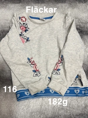 Vit sweatshirt med broderade blommor - Vit sweatshirt från Polarn O. Pyret med broderade blommor i rosa och blått på axel och ärm. Tröjan har långa ärmar och blå ribbad kant nertill med logga. Perfekt för en avslappnad och söt stil.🌸 Fläckar. Storlek 116