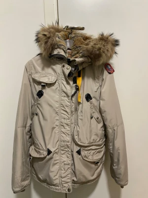 Beige dunjacka från Parajumpers - Snygg beige dunjacka från Parajumpers med stor huva och äkta pälsdetalj. Jackan har flera fickor, dragkedja och knappar framtill samt en gul rem vid kragen. Perfekt för kalla dagar och riktigt varm. Jackan kostar igentligen 10 00kr, men priset jag säljer den för kan diskuteras! 