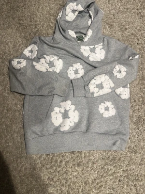 Grå hoodie från Denim Tears med blommor - Säljer en grå hoodie från Denim Tears med vita blommönster över hela tröjan. Hoodien har en stor ficka framtill och en klassisk huva. Tillverkad i mjukt material som känns skönt mot huden. Perfekt för dig som gillar streetwear och unika prints.