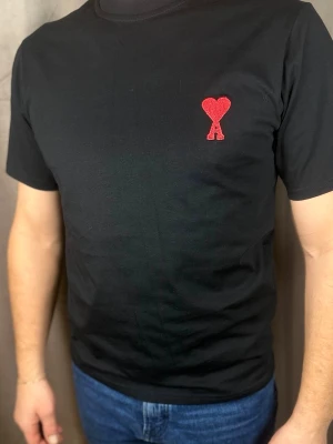 Svart t-shirt från Ami Paris - Svart t-shirt från Ami Paris med röd broderad logga i form av ett hjärta och bokstaven A på bröstet. Klassisk rund hals och korta ärmar. Tillverkad i mjuk bomull för en skön känsla.