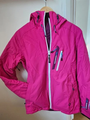 Rosa skidjacka från Tenson - Vadderad skidjacka från Tenson med huva, vit dragkedja och otroligt många praktiska fickor med dragkedja. Storlek 36. Eller motsvarande junior 170 cl. Ficka på ärmen för liftkort. Vädringsdragkedjor i armhålorna. Snölås i midjan. Muddar med hål för tumme ärmslut. Jackan har justerbar nederkant och kontrasterande detaljer. Figursydd. Perfekt för blåsiga dagar och outdoor-aktiviteter. Använd en gång. 